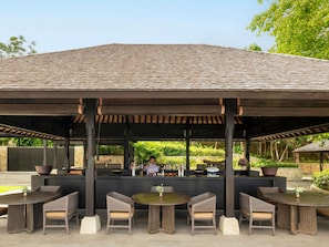 An outdoor seating area with tables and chairs, a covered structure, and a bar area with a person behind the counter. - Raffles Bali (Jimbaran)
