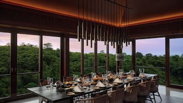 Un espace repas avec une longue table dressée pour le repas, de grandes fenêtres offrant une vue sur la forêt et des lampes suspendues.