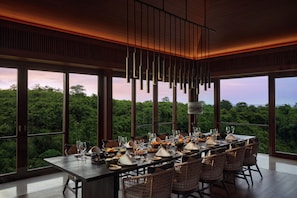 A dining area with a long table set for a meal, large windows offering a view of the forest, and hanging pendant lights. - Raffles Bali (Jimbaran)