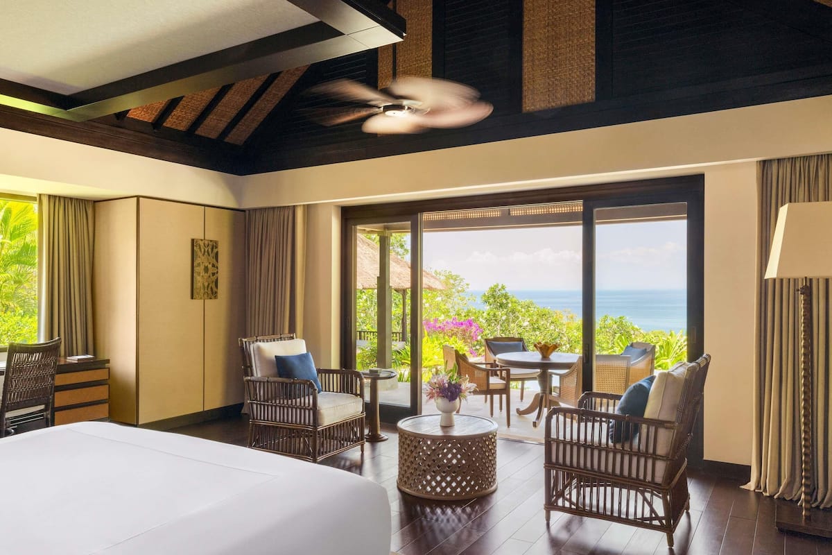 A hotel room with a bed, wicker chairs, a round table, and a view of the ocean.