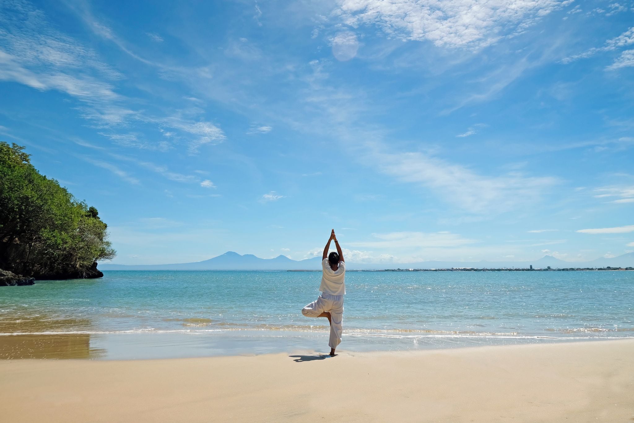 Eine Person führt eine Yoga-Pose am Strand aus, im Hintergrund ein klarer Himmel und Berge.