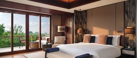 A spacious bedroom with a large bed, a sitting area with a chair and table, and a balcony with a view of greenery.