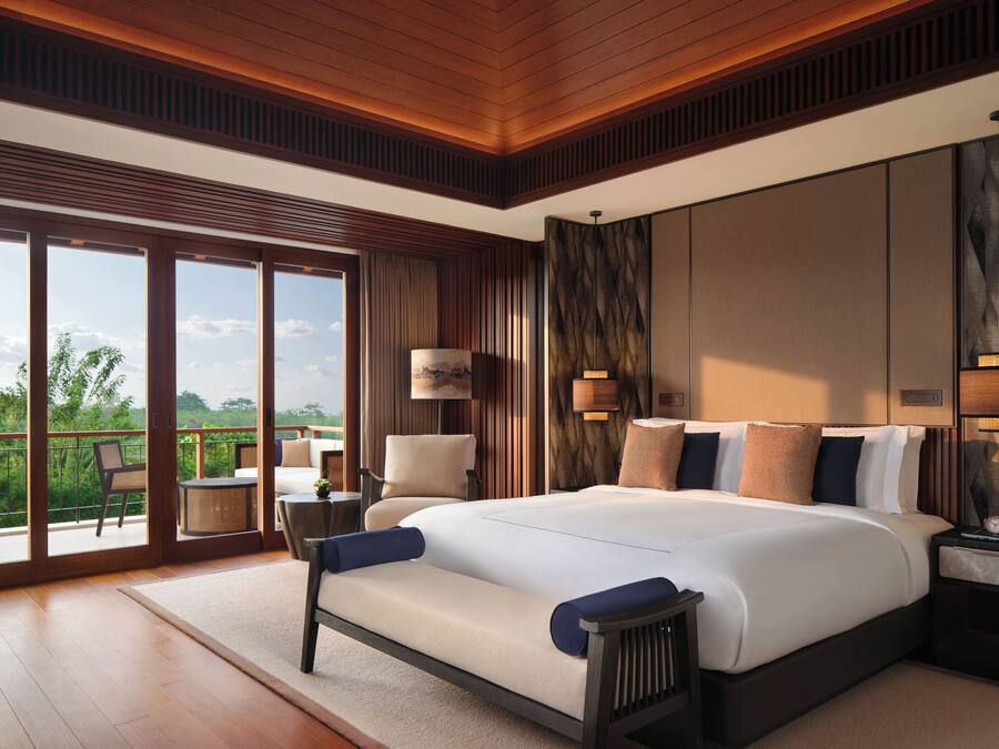 A spacious bedroom with a large bed, a sitting area with a chair and table, and a balcony with a view of greenery.