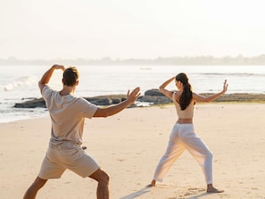 Two people practicing Tai Chi on a beach with a view of the ocean and rocks in the background. - Raffles Bali (Jimbaran)
