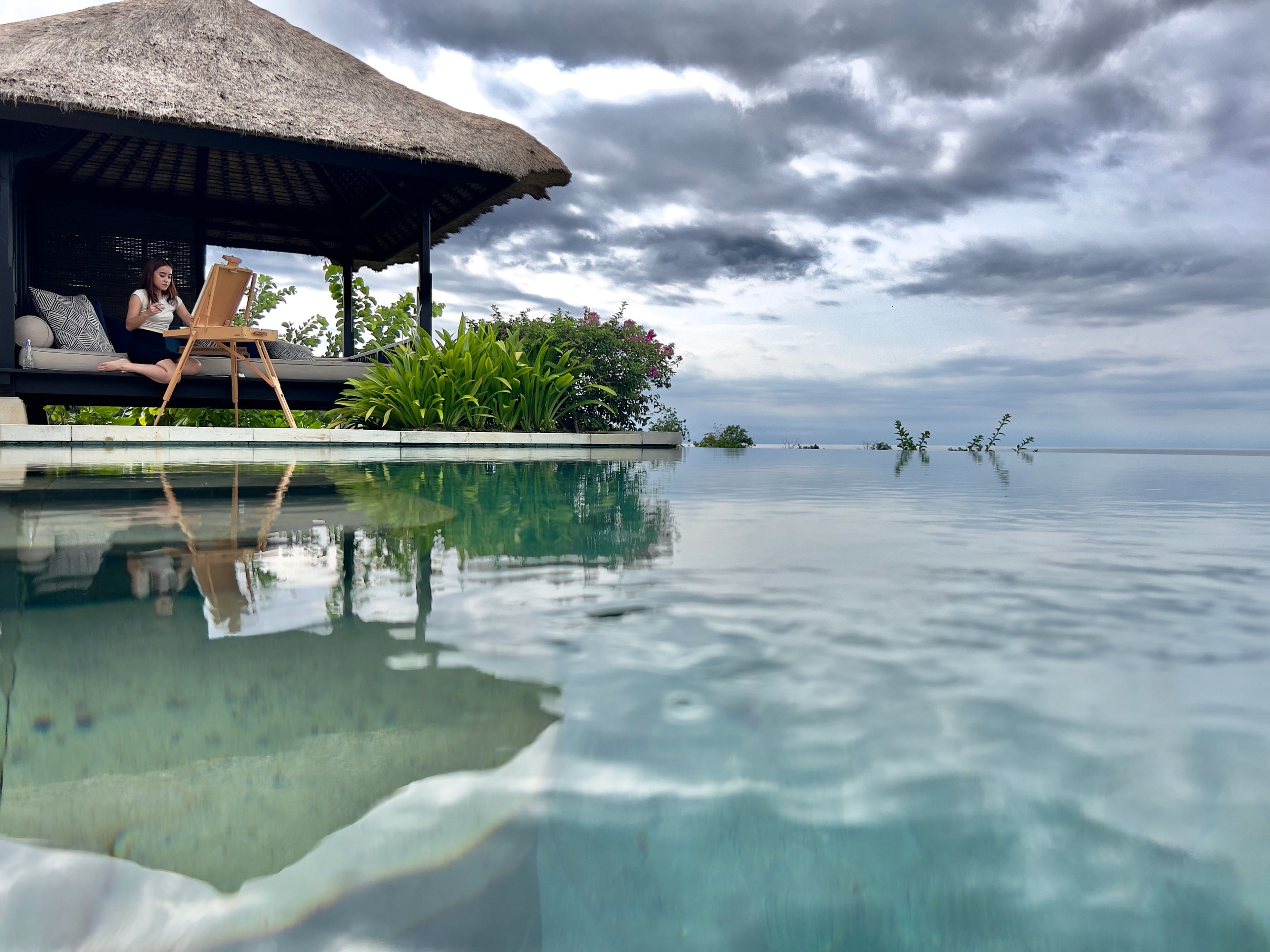 Una piscina infinita tranquila con vista despejada al océano, una estructura con techo de paja y exuberante vegetación.