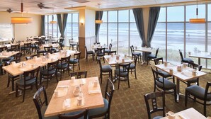 A spacious dining area with tables and chairs, large windows with curtains, and a view of the beach. - Myrtle Beach | Family Beach Retreat | Pool + Beach | 2BR (Myrtle Beach)