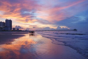 A beach with a pier extending into the water, a group of people walking along the shore, and buildings in the background.