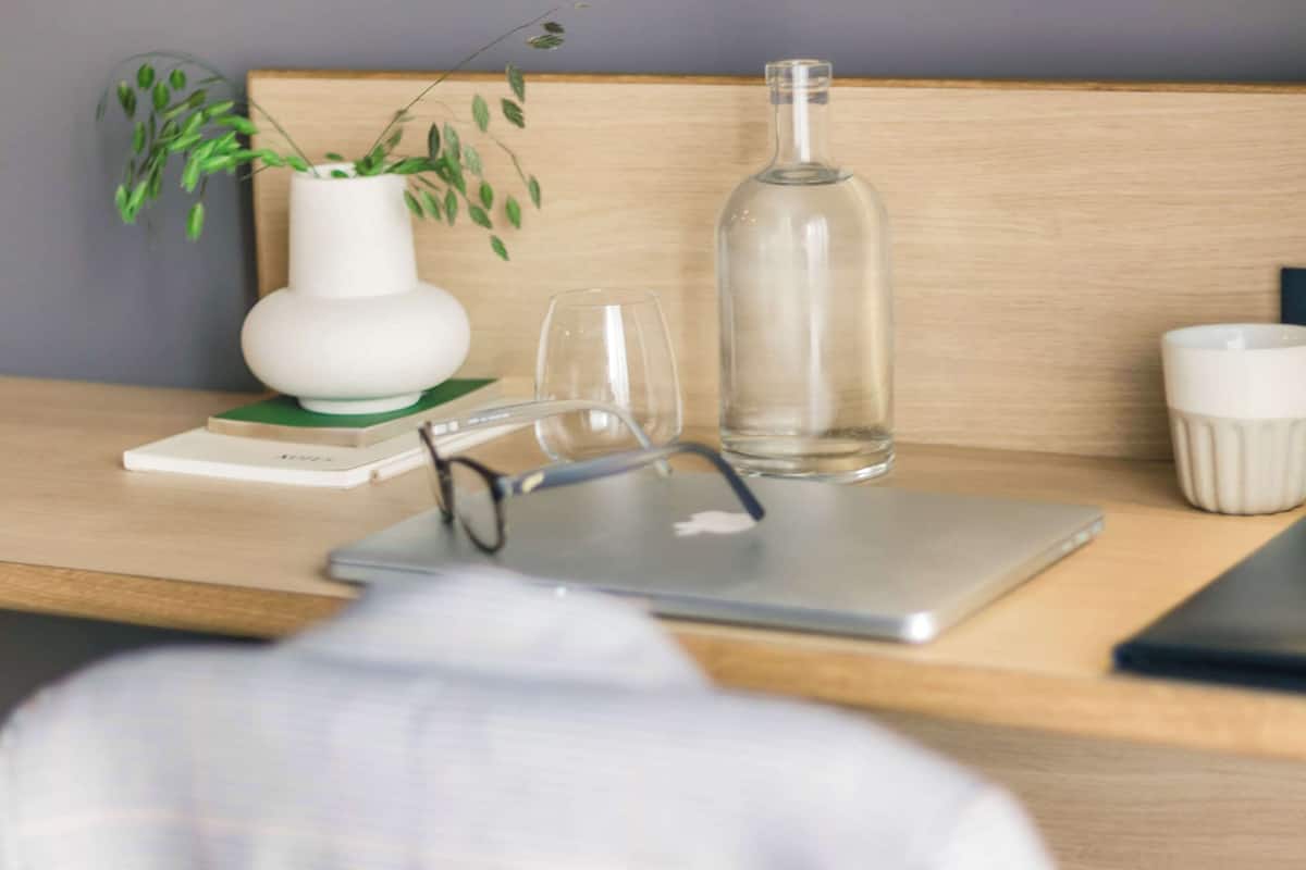 Un bureau en bois avec un ordinateur portable, une bouteille en verre transparent, un vase blanc avec une plante, une paire de lunettes et une tasse blanche.