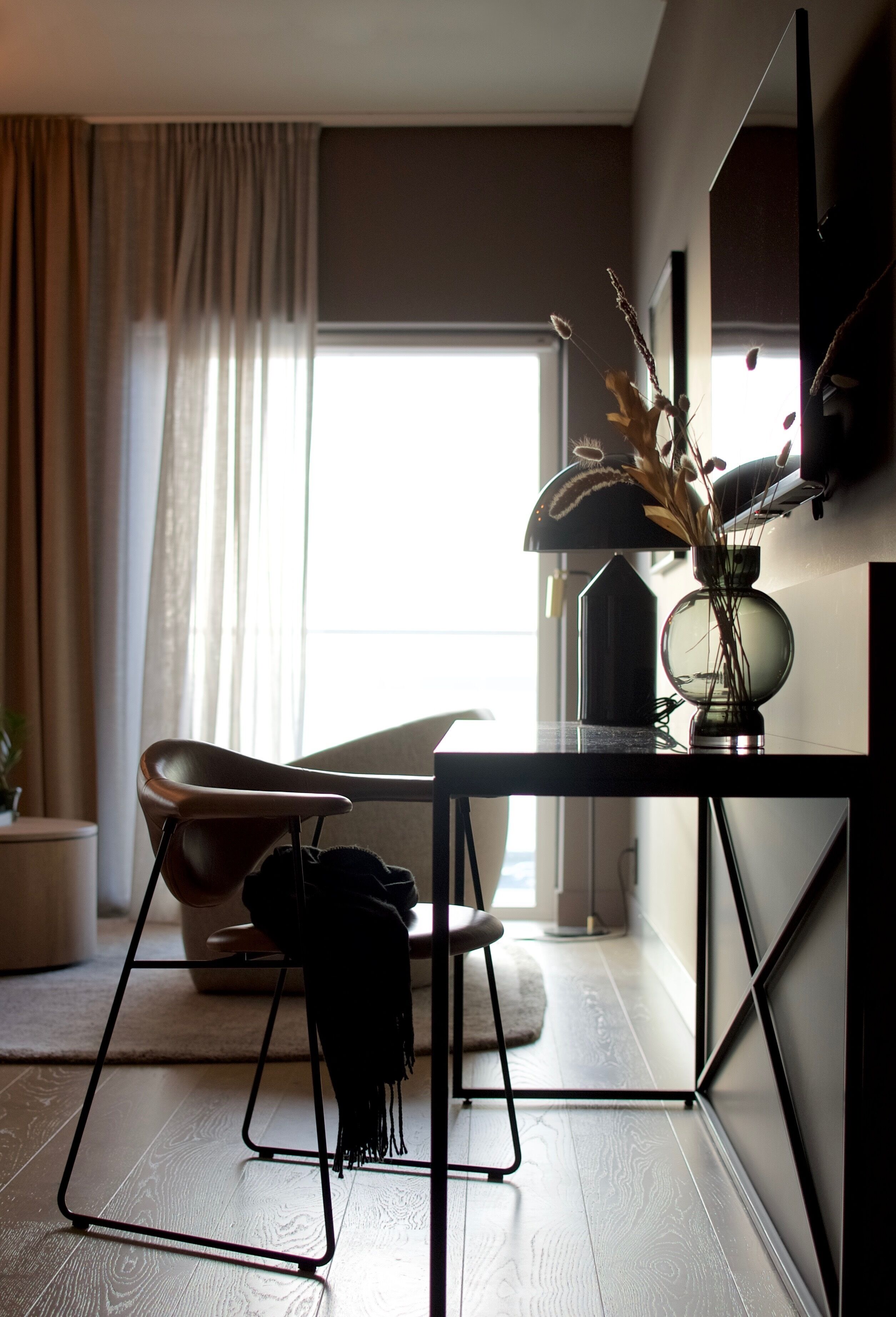 A modern living room with a minimalist design, featuring a black table, a leather chair, and a vase with dried plants.