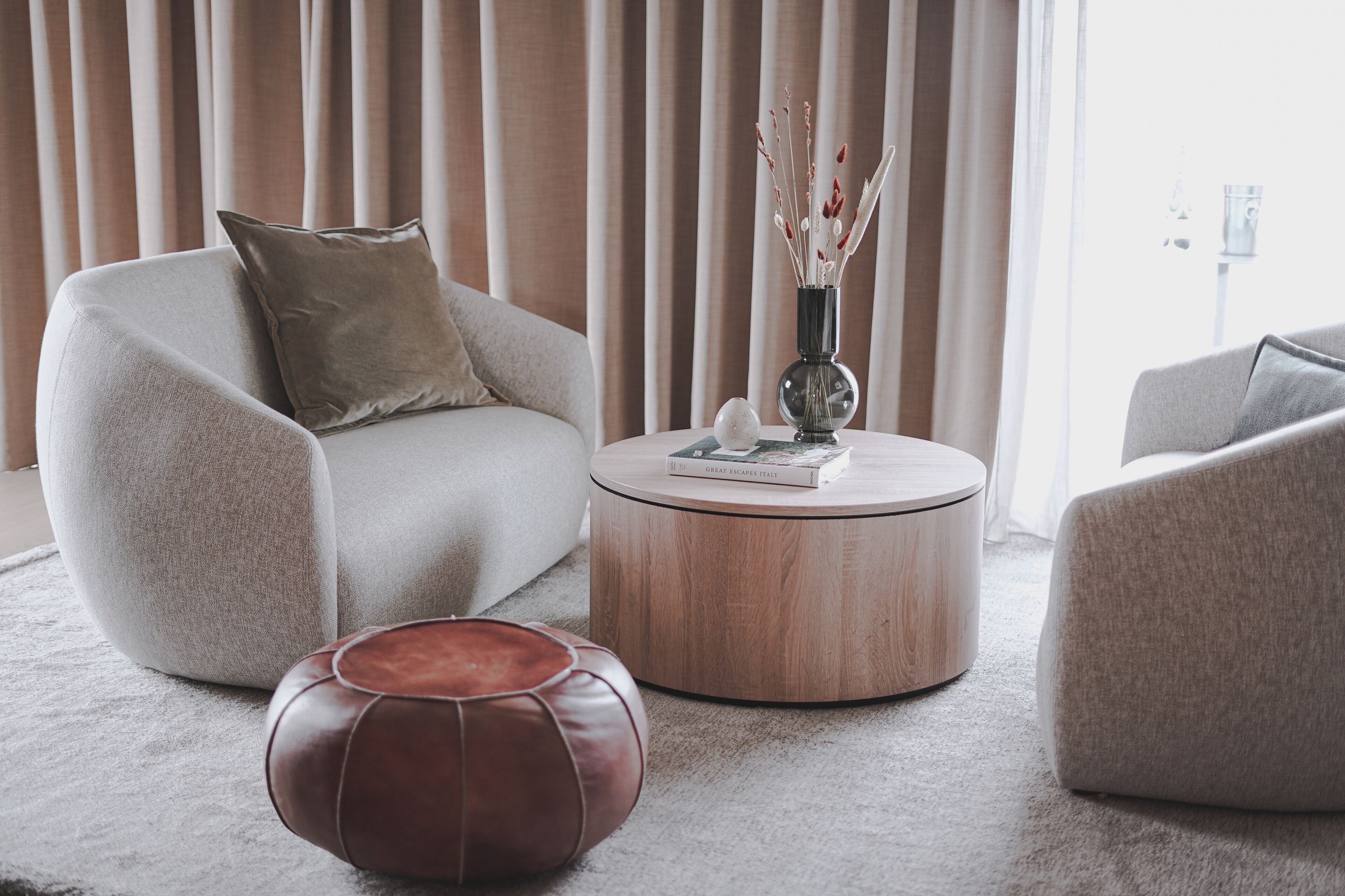 A modern living room with a round wooden coffee table, two grey sofas, a brown leather ottoman, and a vase with dried plants.