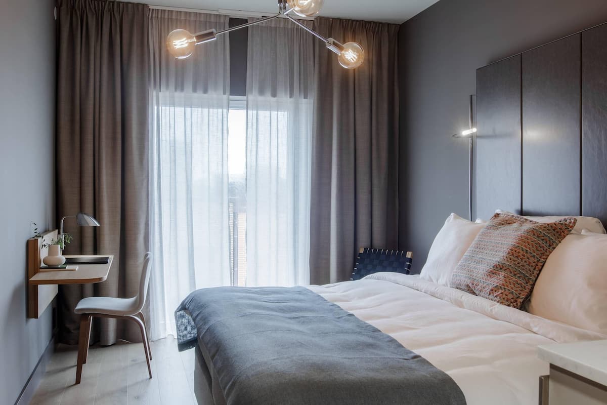 Une chambre moderne équipée d’un lit, d’un bureau et d’une chaise, et offrant une vue sur la ville grâce à de grandes fenêtres.