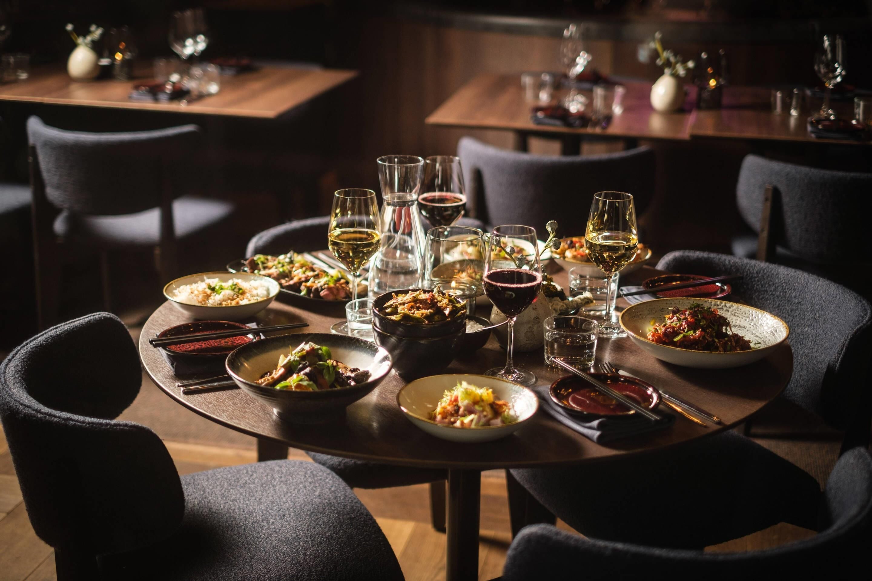 Una mesa de comedor dispuesta con varios platos y copas de vino, rodeada de sillas.