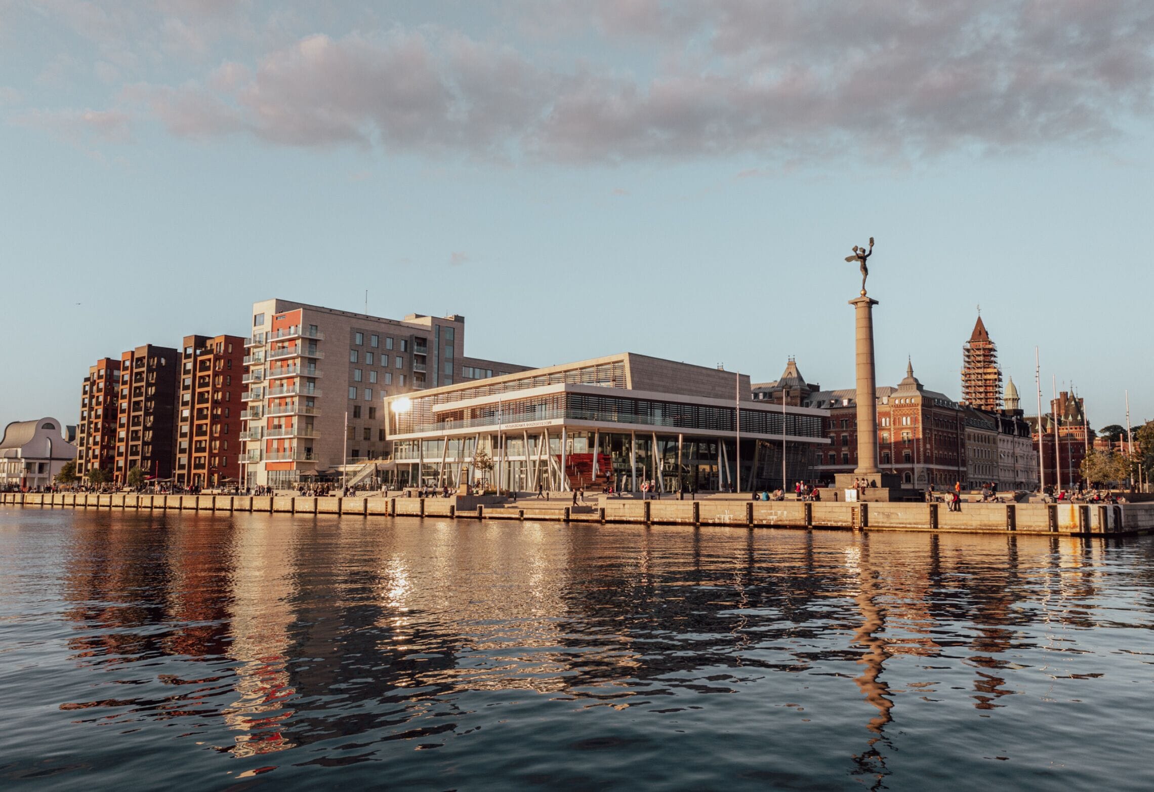 En stadsbild vid vattnet med moderna byggnader, ett markant torn och en stilla flod som speglar byggnaderna.