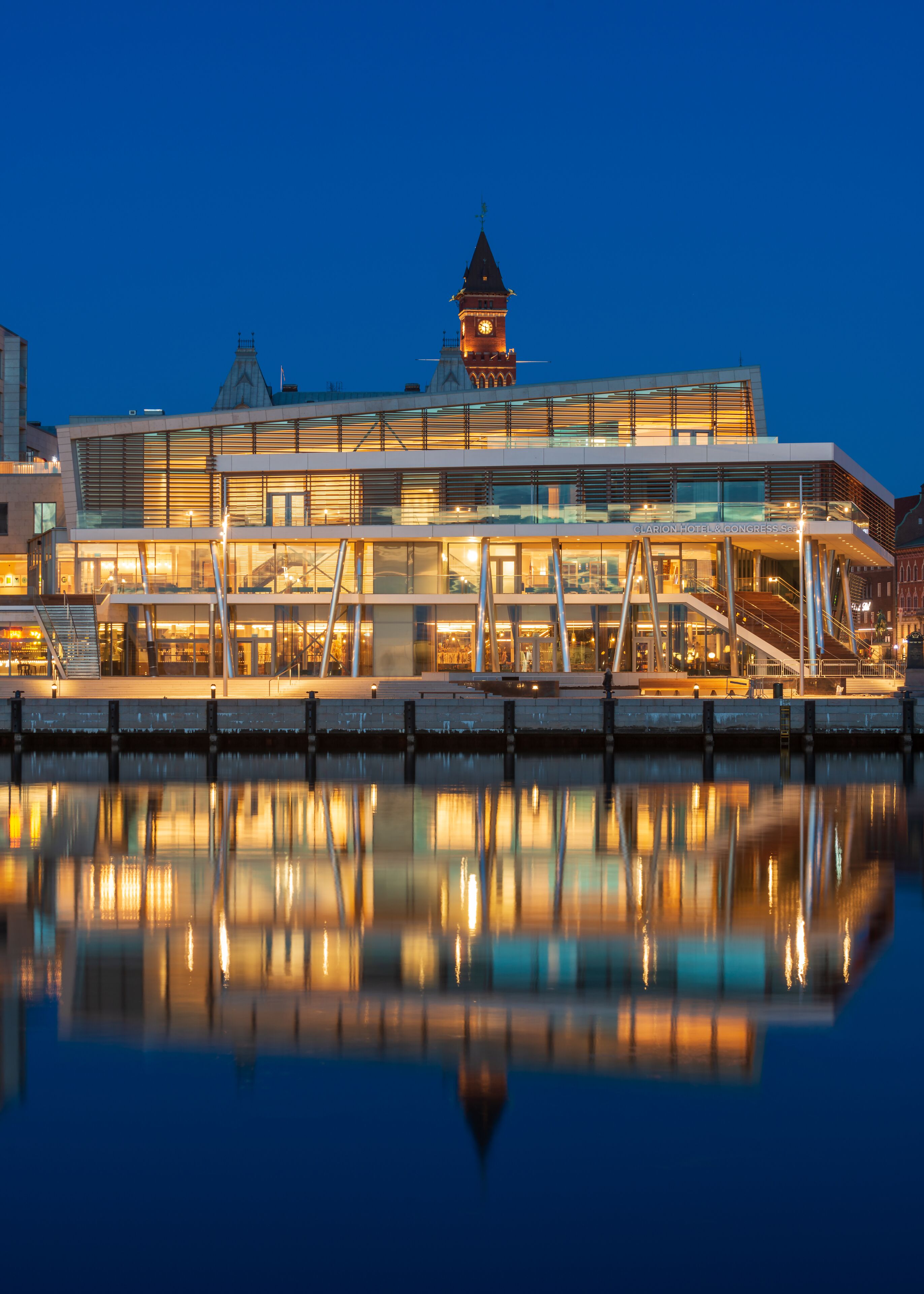 Un hôtel moderne avec une façade en verre, une tour horlogée et son reflet dans l’eau.