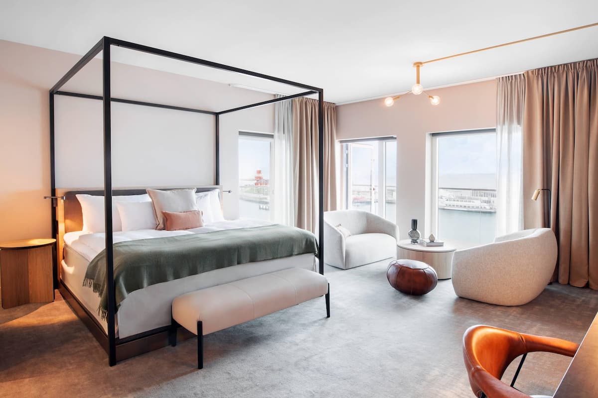 Un lit à baldaquin avec un banc, un fauteuil blanc, un pouf marron et une chaise moderne, dans une chambre d’hôtel dotée de grandes fenêtres et offrant une vue sur la ville.