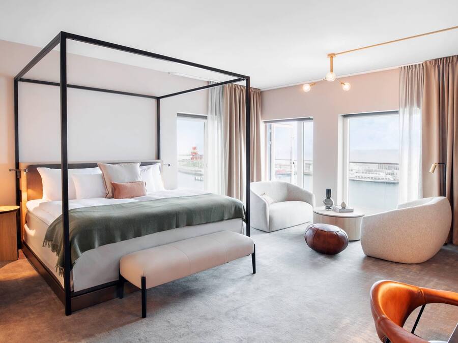 A four-poster bed with a bench, a white armchair, a brown ottoman, and a modern chair in a hotel room with large windows and a city view.