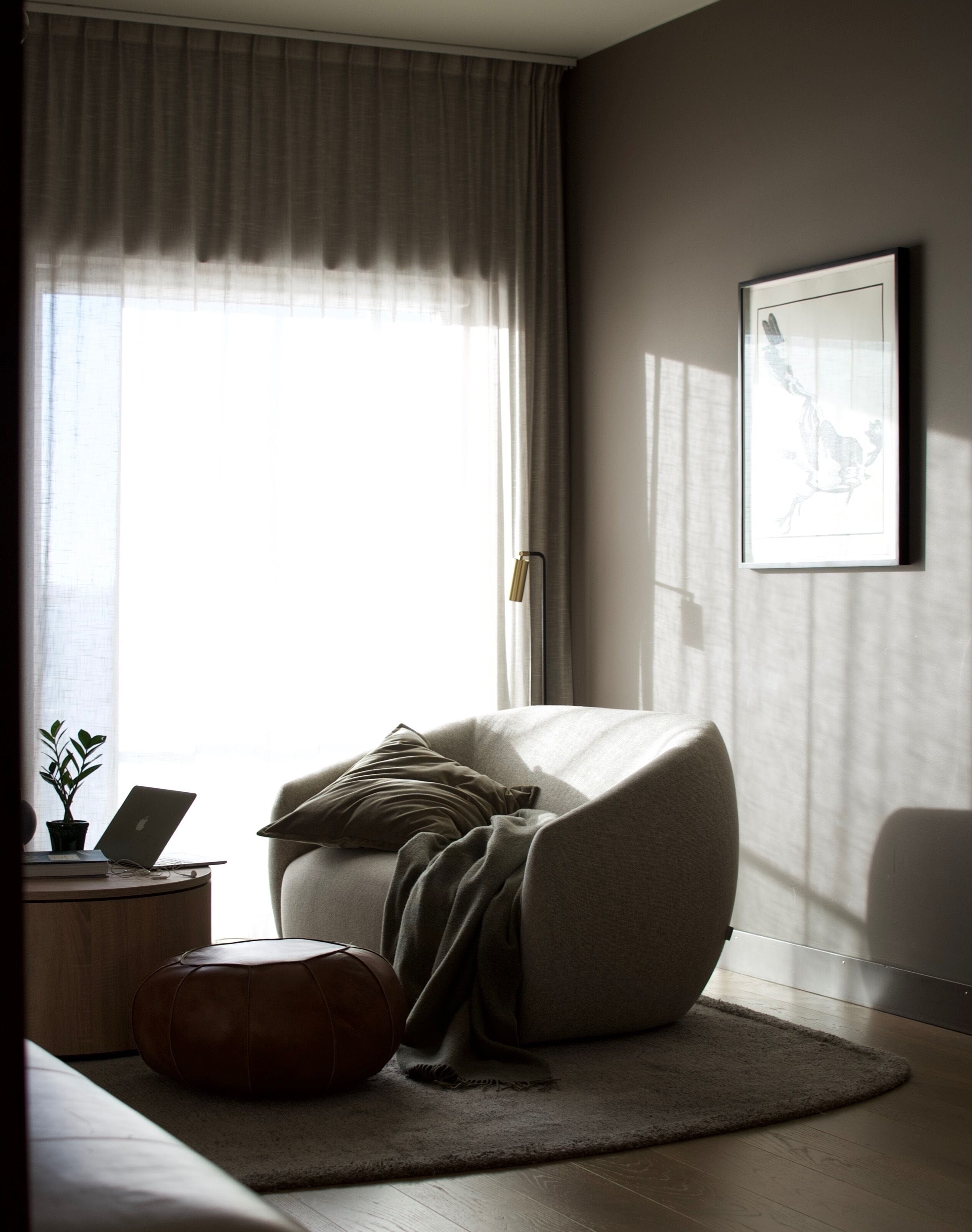 A modern bedroom with a large window, a comfortable chair, a bedside table with a laptop, and a framed picture on the wall.