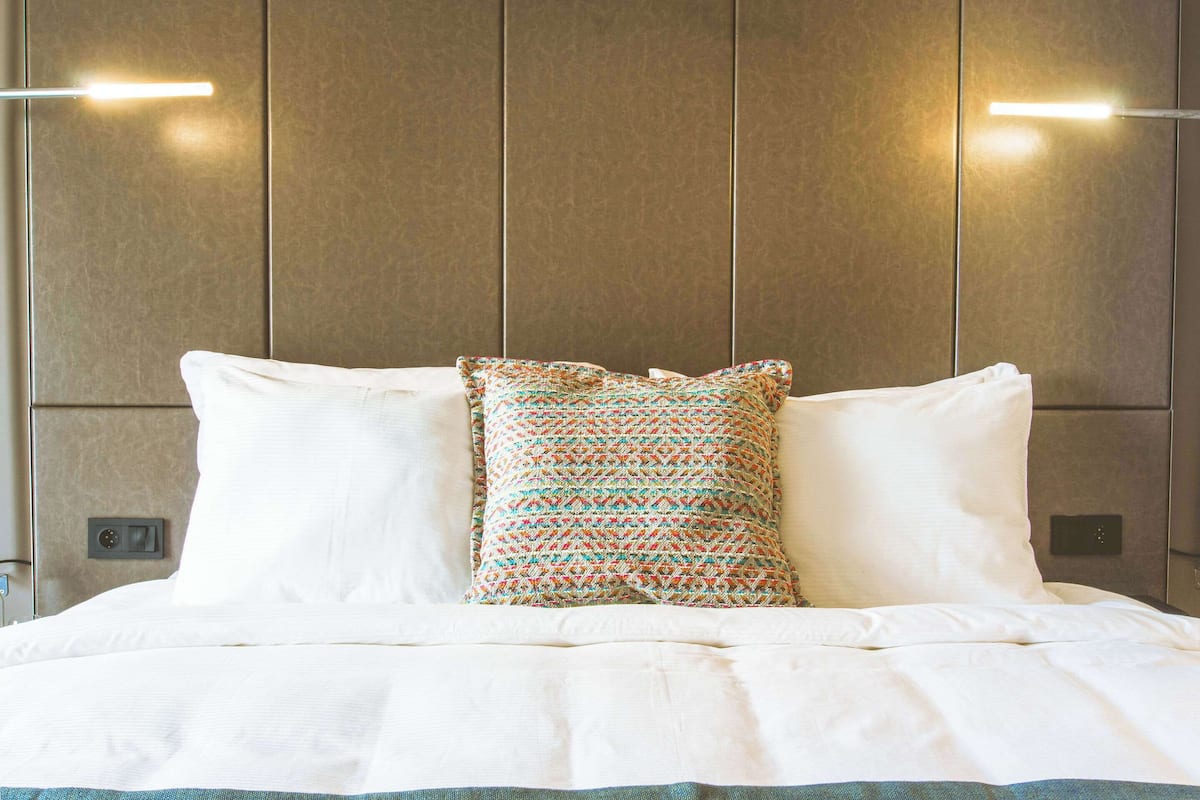 Un lit bien fait, avec du linge de lit blanc et un coussin aux motifs colorés, placé devant un mur orné de lambris verticaux et agrémenté de deux lampes murales.