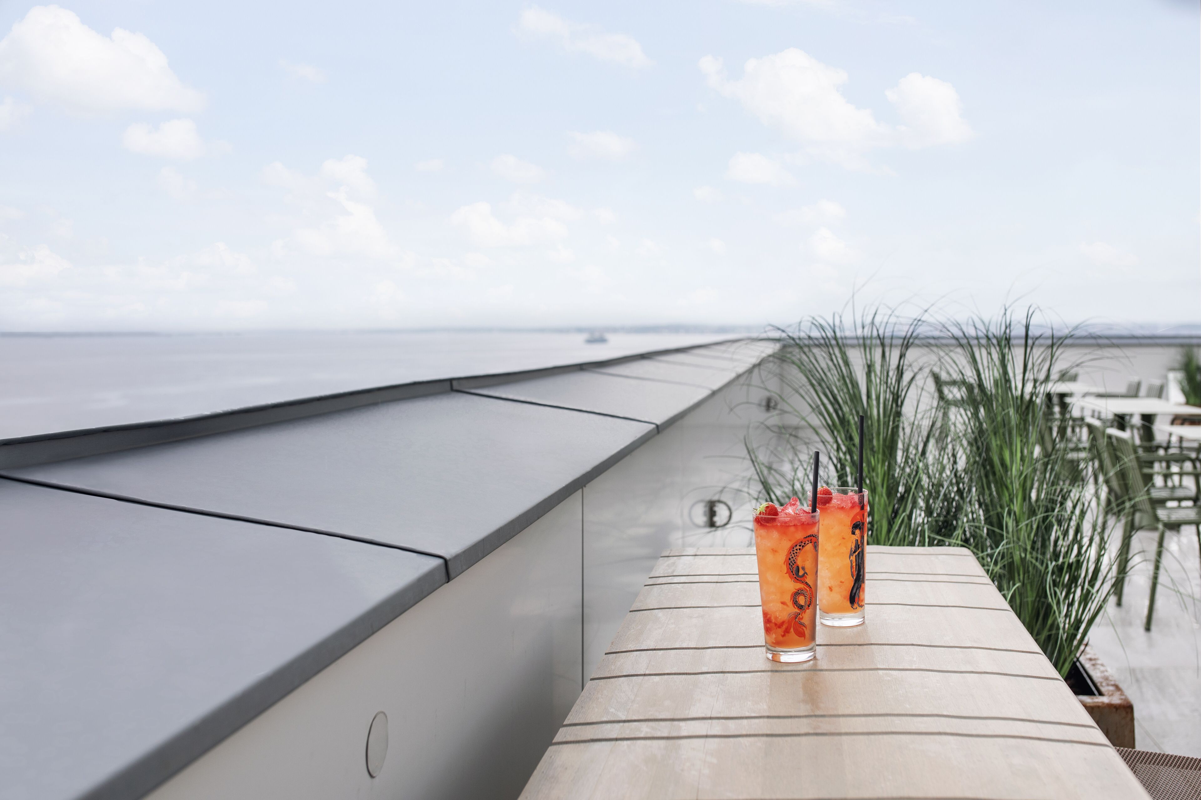 A wooden table with two glasses of iced drinks, a view of the ocean, and some greenery.