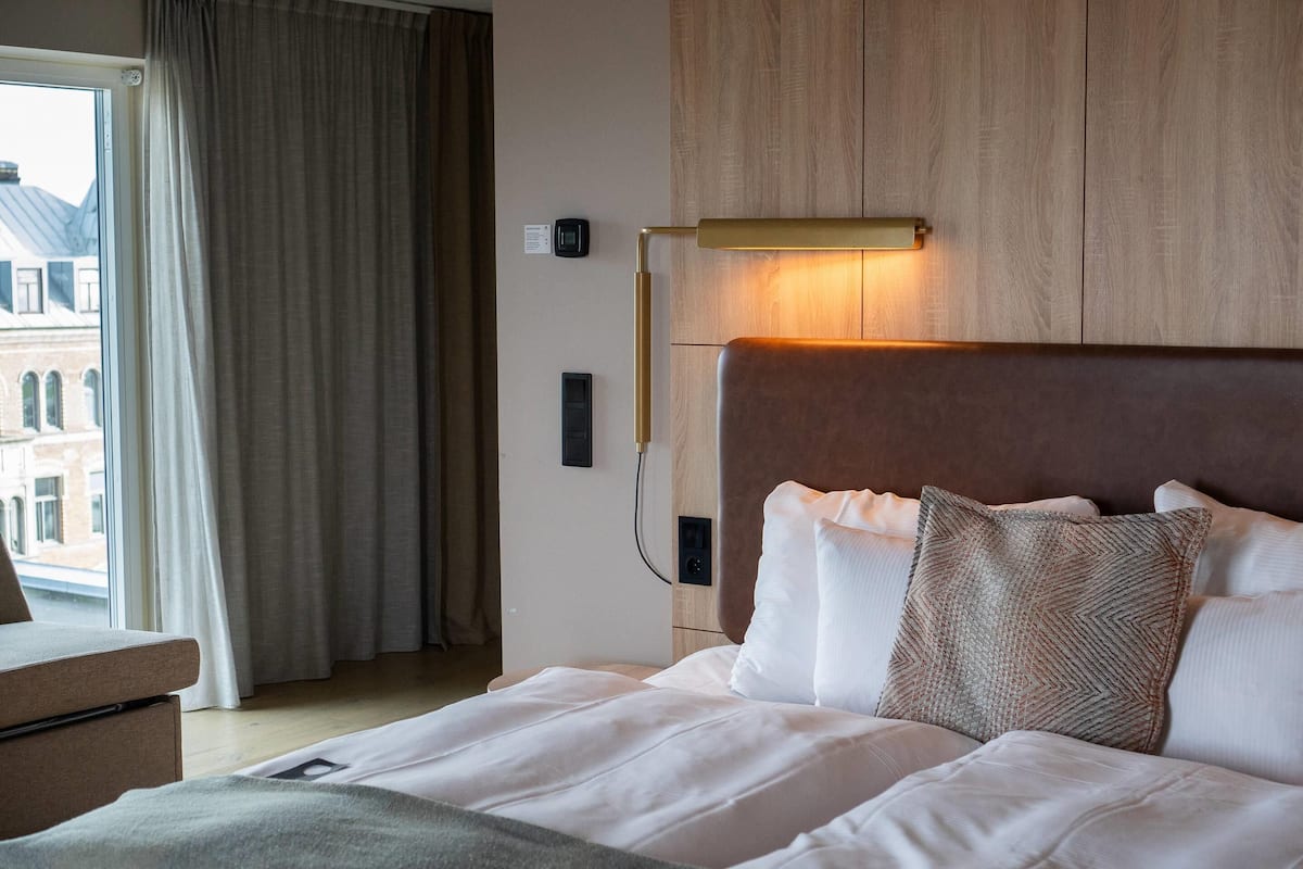 Une chambre d’hôtel avec un lit, une table de chevet, une lampe et une vue sur les bâtiments par la fenêtre.