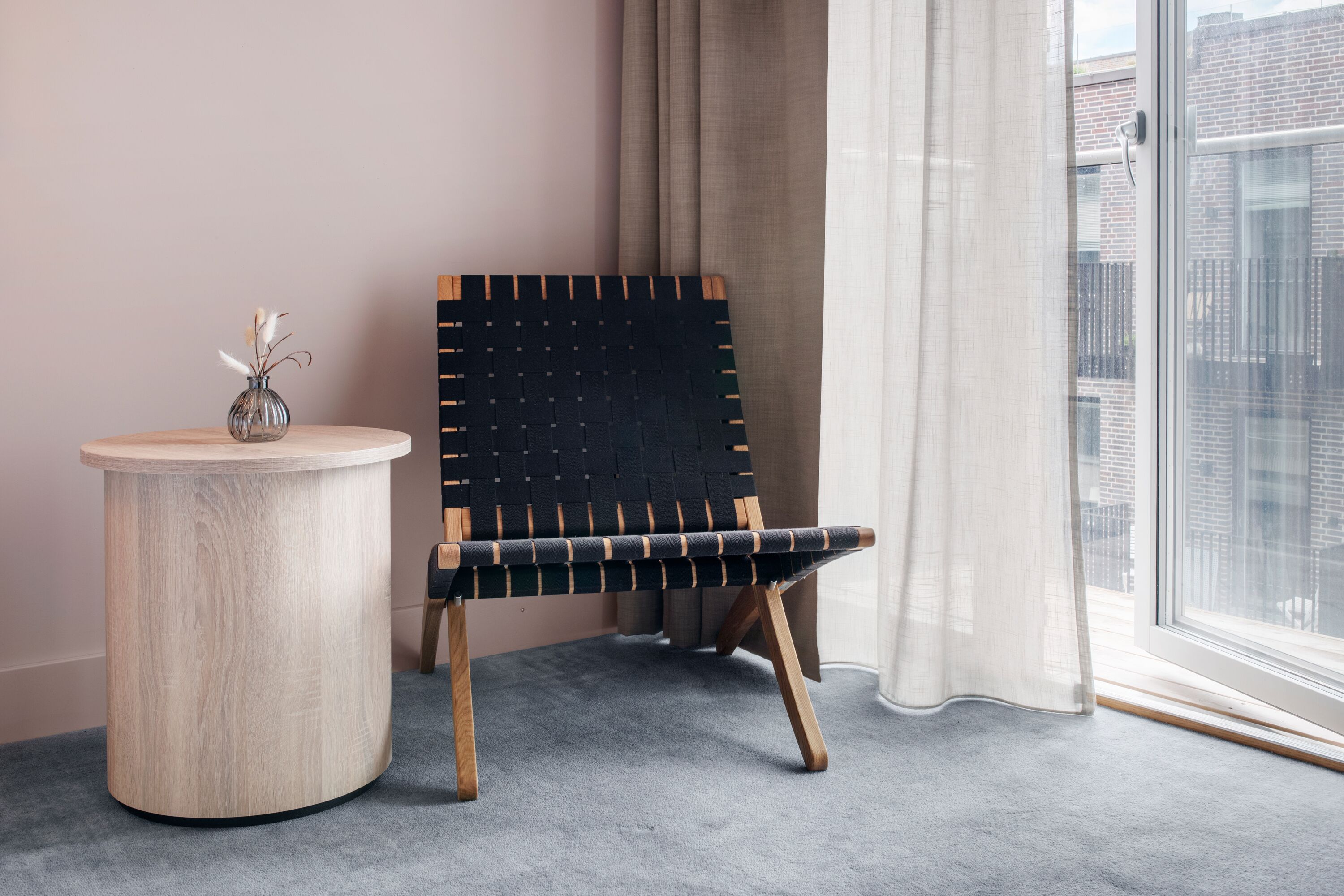 Una habitación con una mesa redonda de madera, una silla negra tejida y vistas a edificios a través de una ventana con cortinas translúcidas.