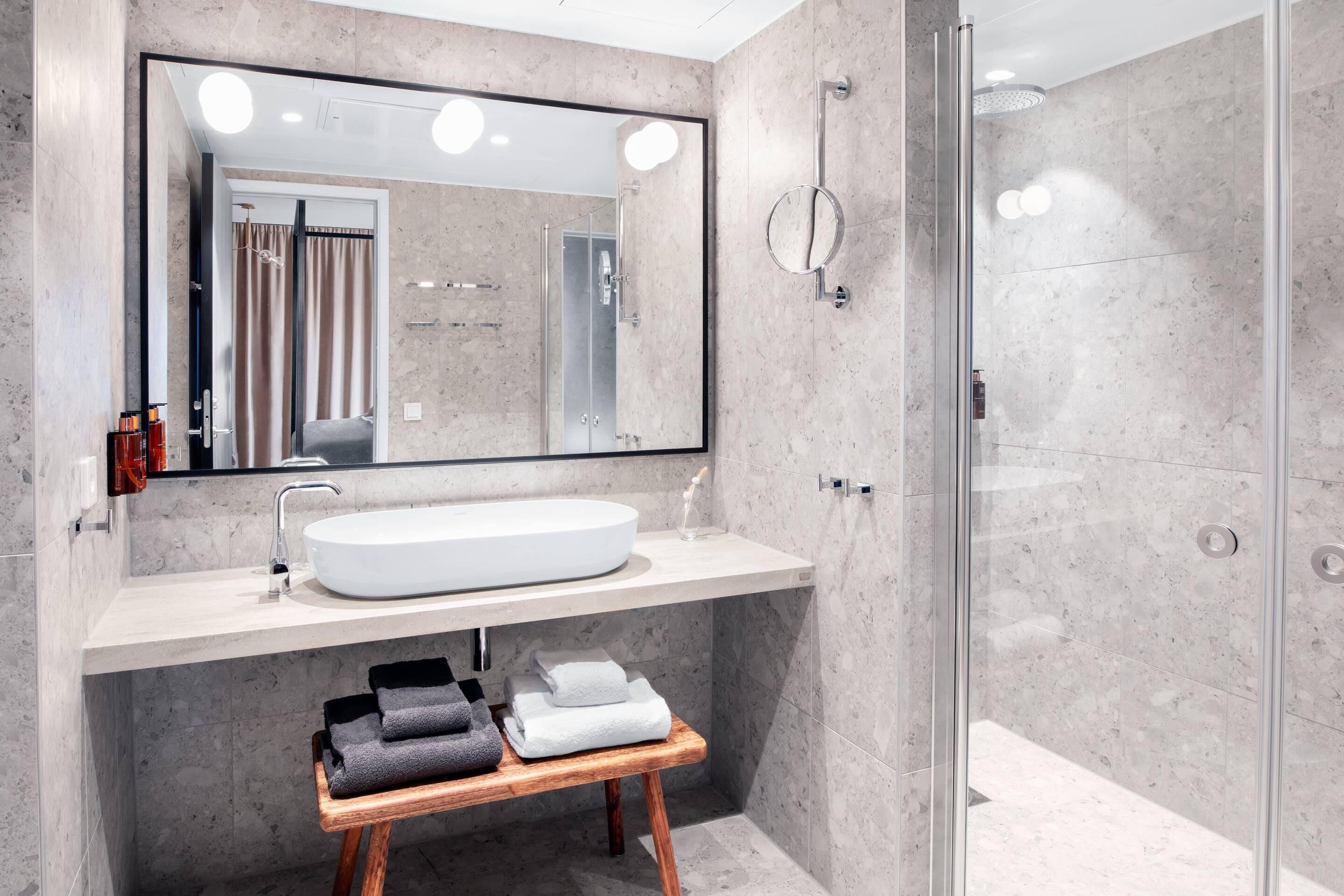 A modern bathroom with a large mirror, a white sink, a wooden stool with towels, and a glass shower enclosure.