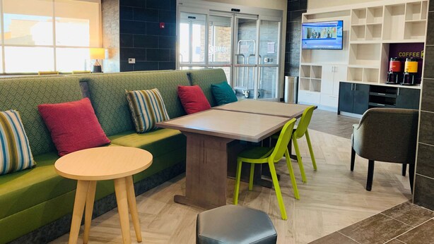 A modern lounge area with a green sofa, a wooden table, and green chairs. There is a coffee station with two coffee machines and a TV on the wall.