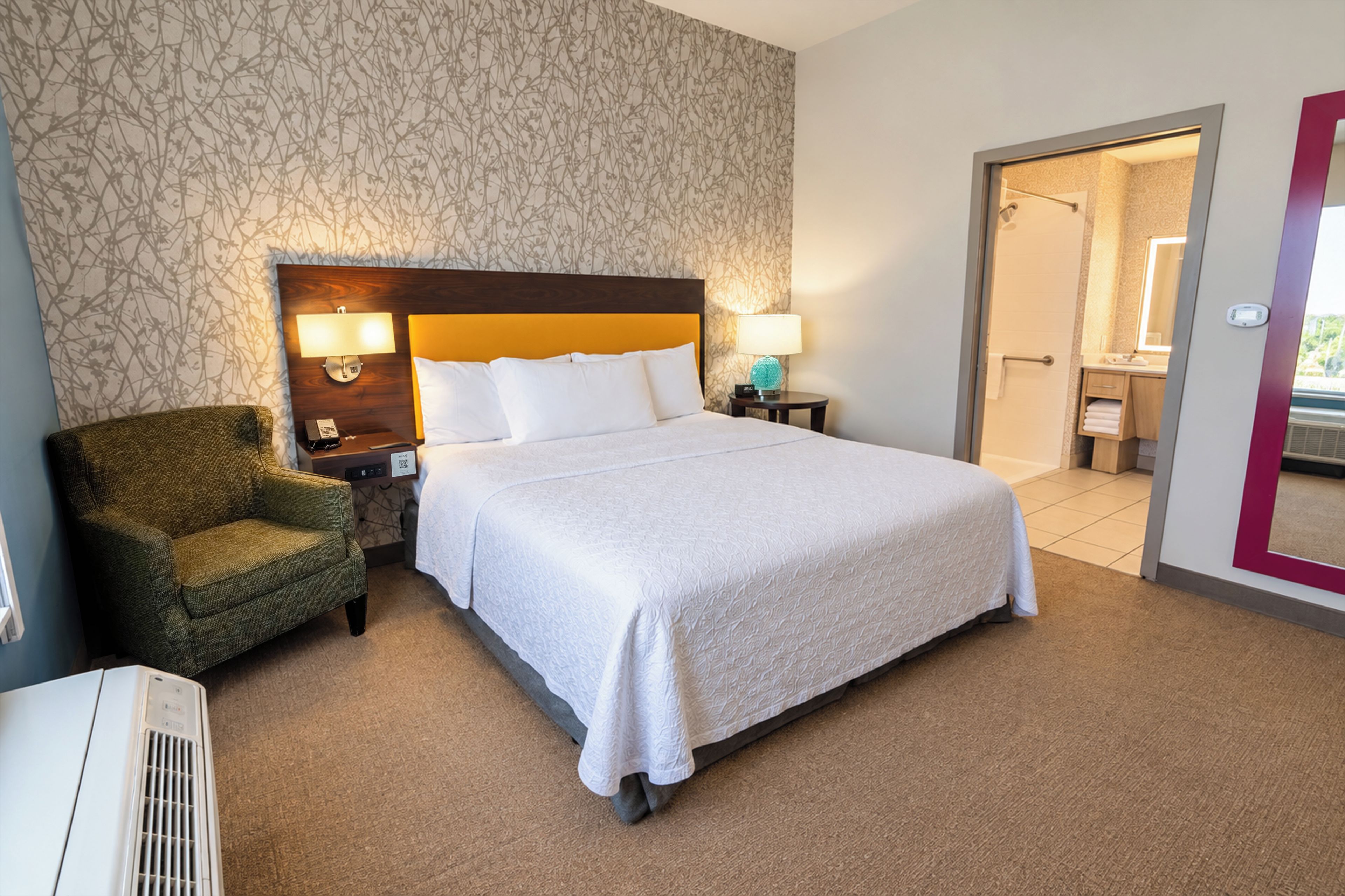 A hotel room with a large bed, a chair, a nightstand, a lamp, and a bathroom visible through an open door.