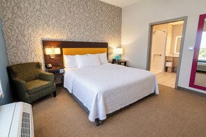 Une chambre d’hôtel avec un grand lit, une chaise, une table de chevet, une lampe et une salle de bain visible à travers une porte ouverte.