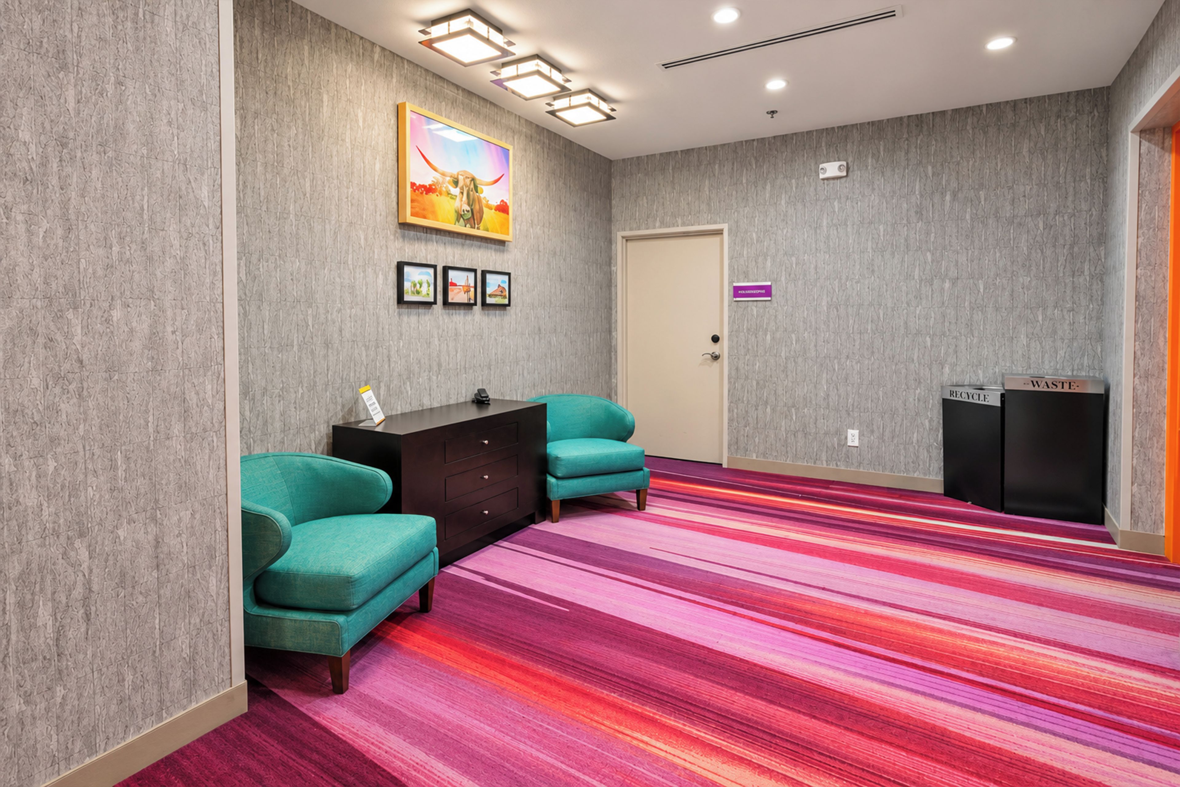 Habitación de hotel con sillones azul verdosos, un escritorio de madera oscura, una alfombra colorida a rayas, una puerta con un letrero morado y una obra de arte colgada en la pared.