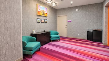 Habitación de hotel con sillones azul verdosos, un escritorio de madera oscura, una alfombra colorida a rayas, una puerta con un letrero morado y una obra de arte colgada en la pared.