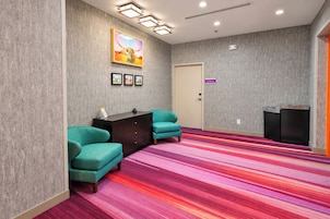 Habitación de hotel con sillones azul verdosos, un escritorio de madera oscura, una alfombra colorida a rayas, una puerta con un letrero morado y una obra de arte colgada en la pared.