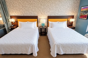 Quarto de hotel com duas camas, uma mesa de cabeceira com abajur, um quadro na parede e papel de parede estampado.