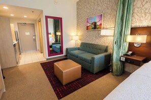 A hotel room with a sofa, ottoman, and a bed. There is a mirror, a painting, and a lamp on the wall. - Home2 Suites by Hilton Edinburg, TX (Edinburg)