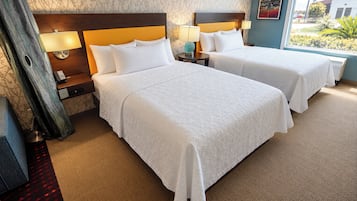 Habitación de hotel con dos camas, una mesita de noche con lámpara, una ventana con vistas y un papel tapiz estampado en la pared.