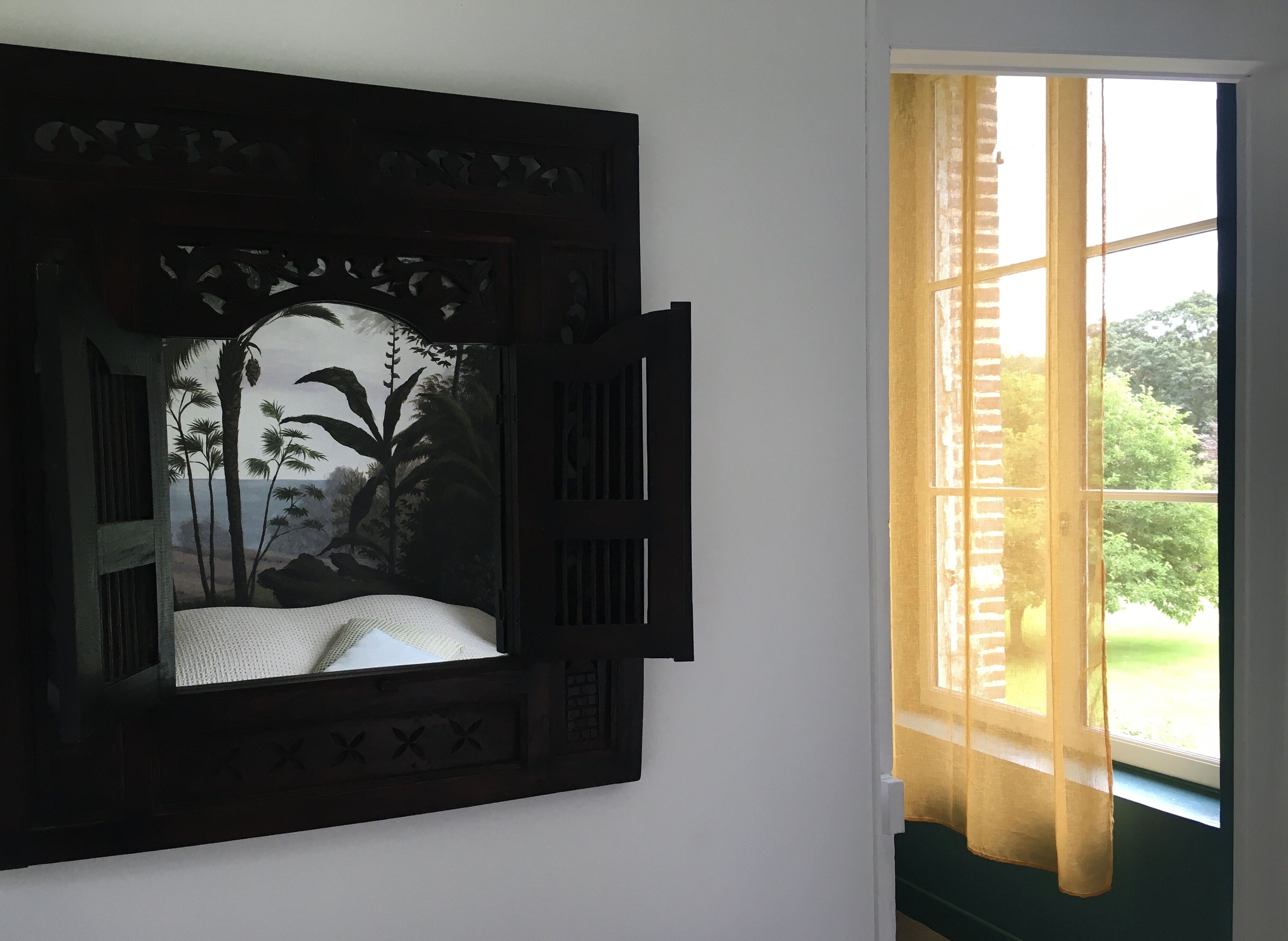 A room with a bed against a wall, a window with curtains, and a view of greenery outside.
