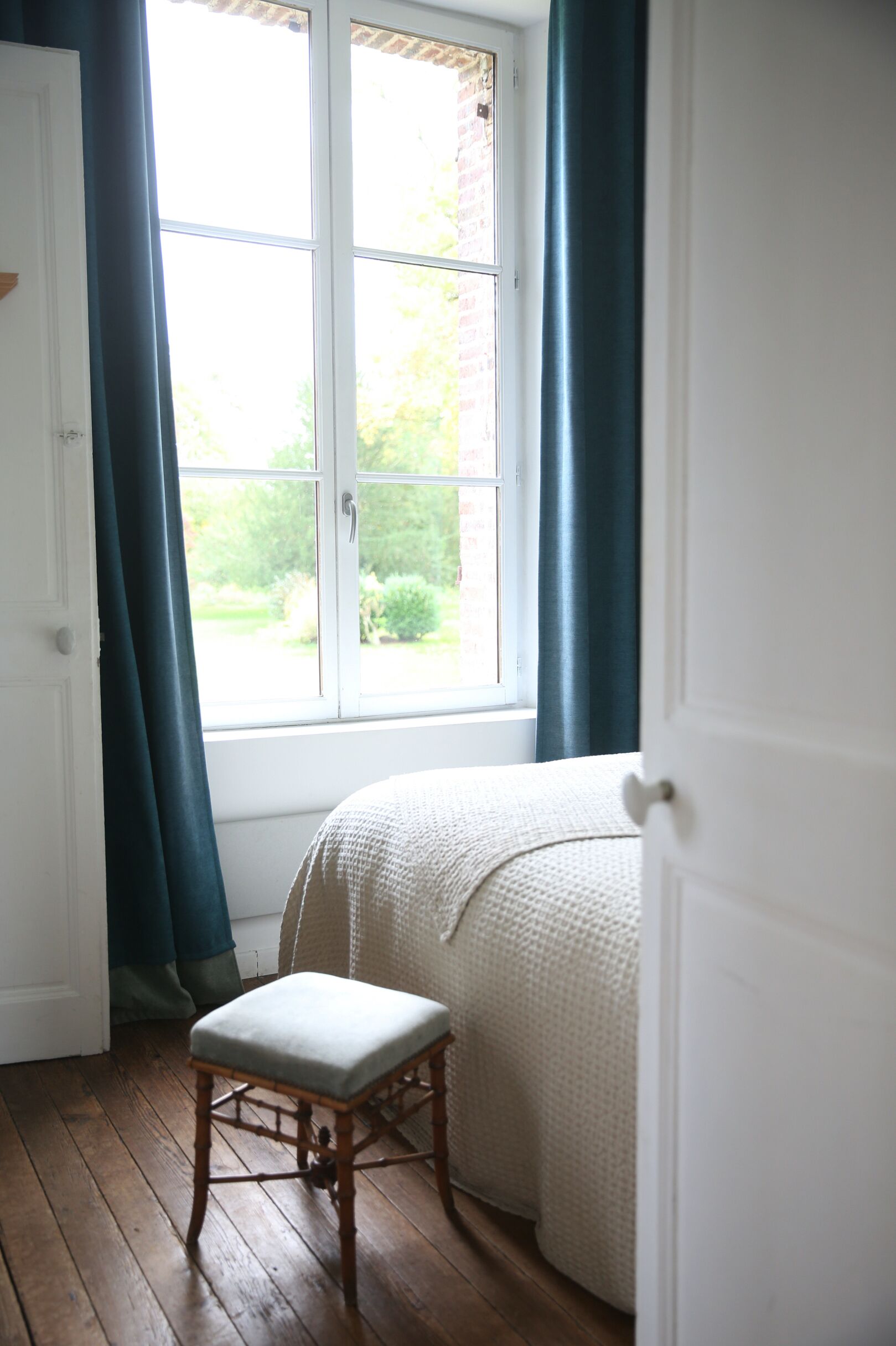 A bedroom with a bed, a window with curtains, a wooden floor, and a bench.