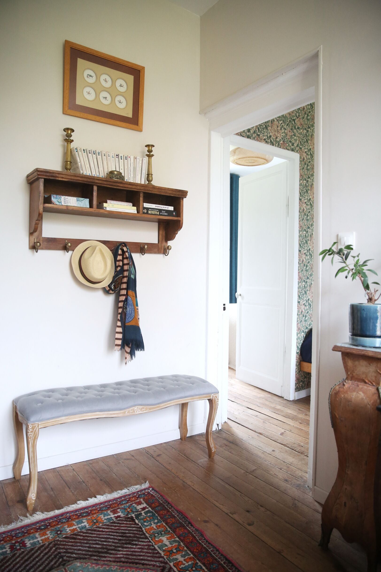 A room with a wooden bench, a hat, and a scarf hanging on the wall, a bookshelf with books, a framed picture, and a potted plant.