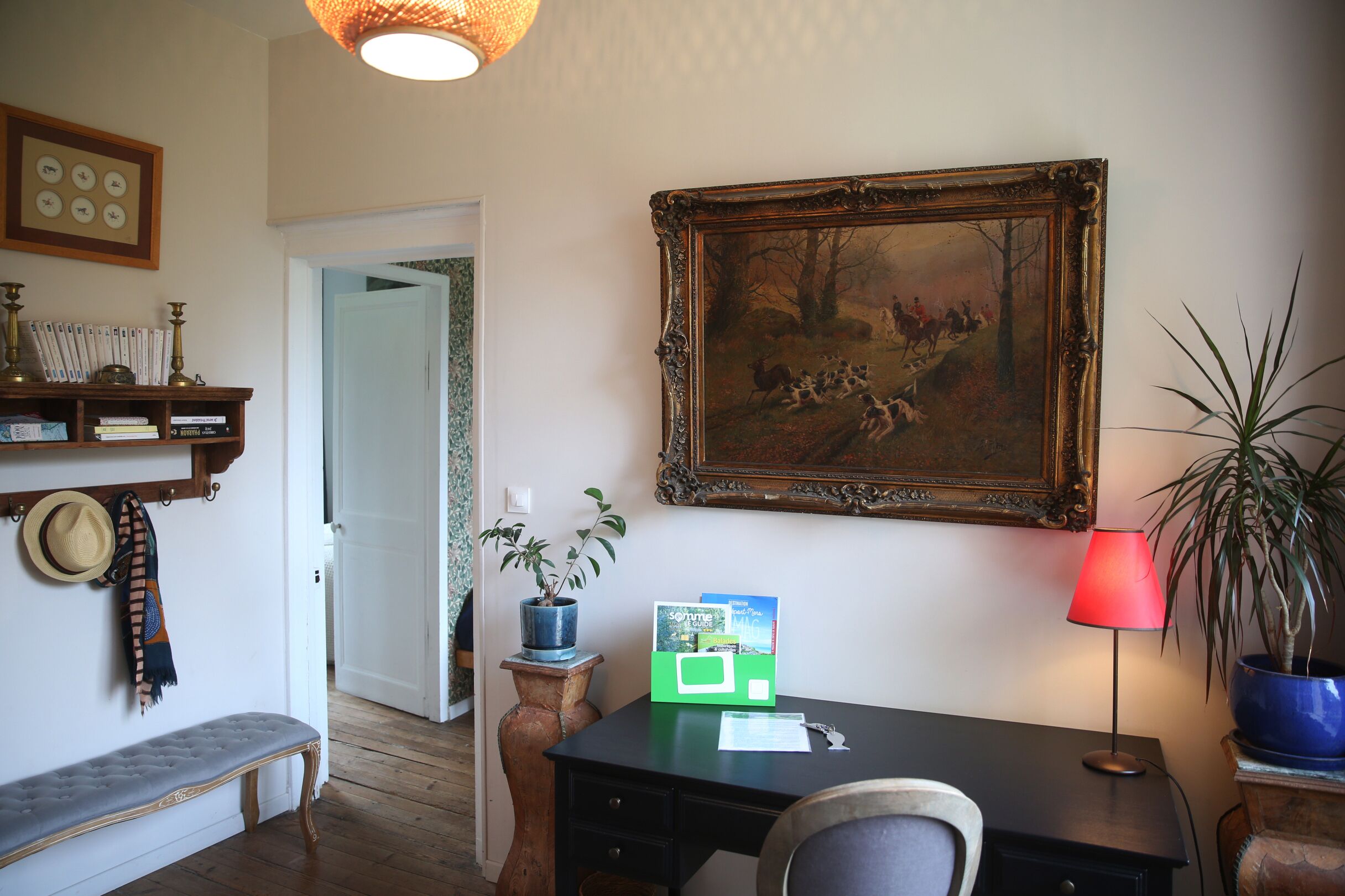A room with a desk, a chair, a bench, a potted plant, a framed painting, and a lamp.