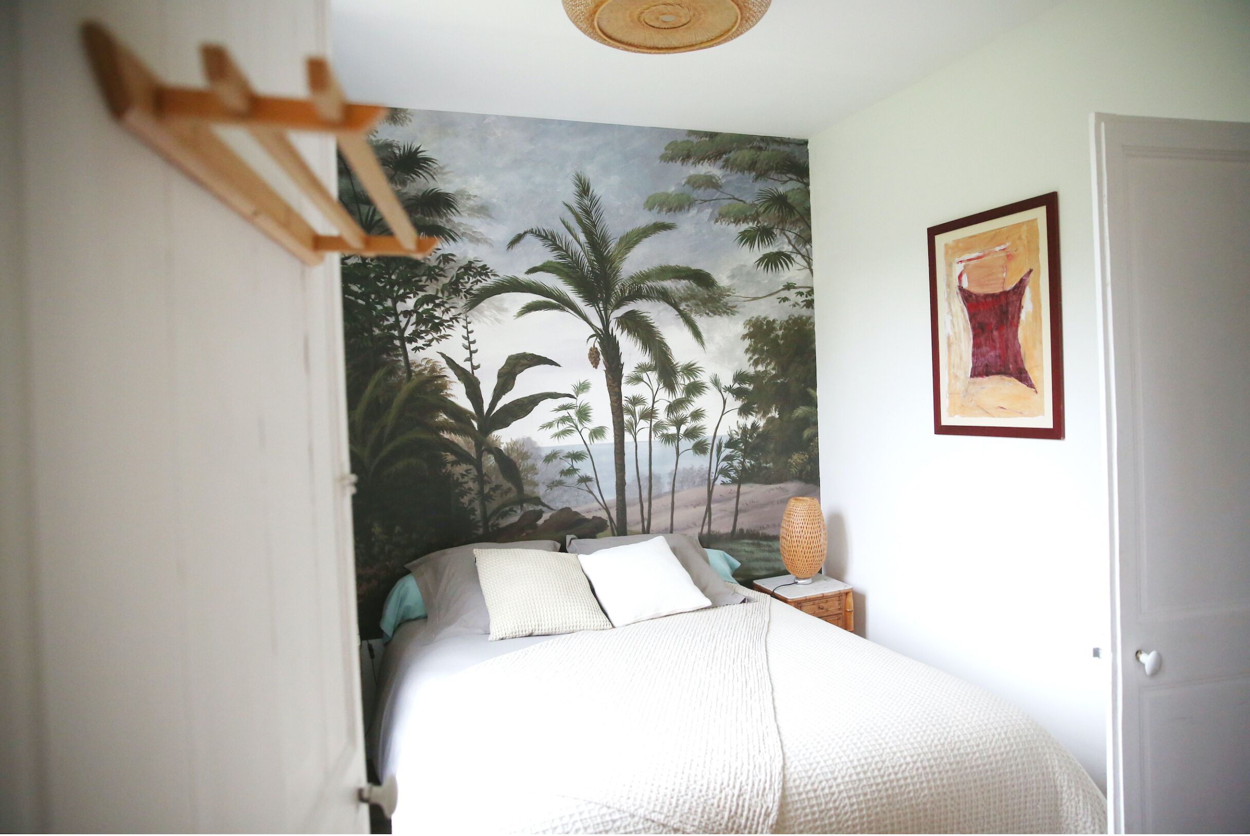 A bedroom with a bed, a nightstand, a wall-mounted lamp, a door, and a painting.