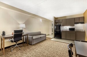 Quarto de hotel compacto com cozinha compacta, sofá, escrivaninha com luminária e uma cadeira.