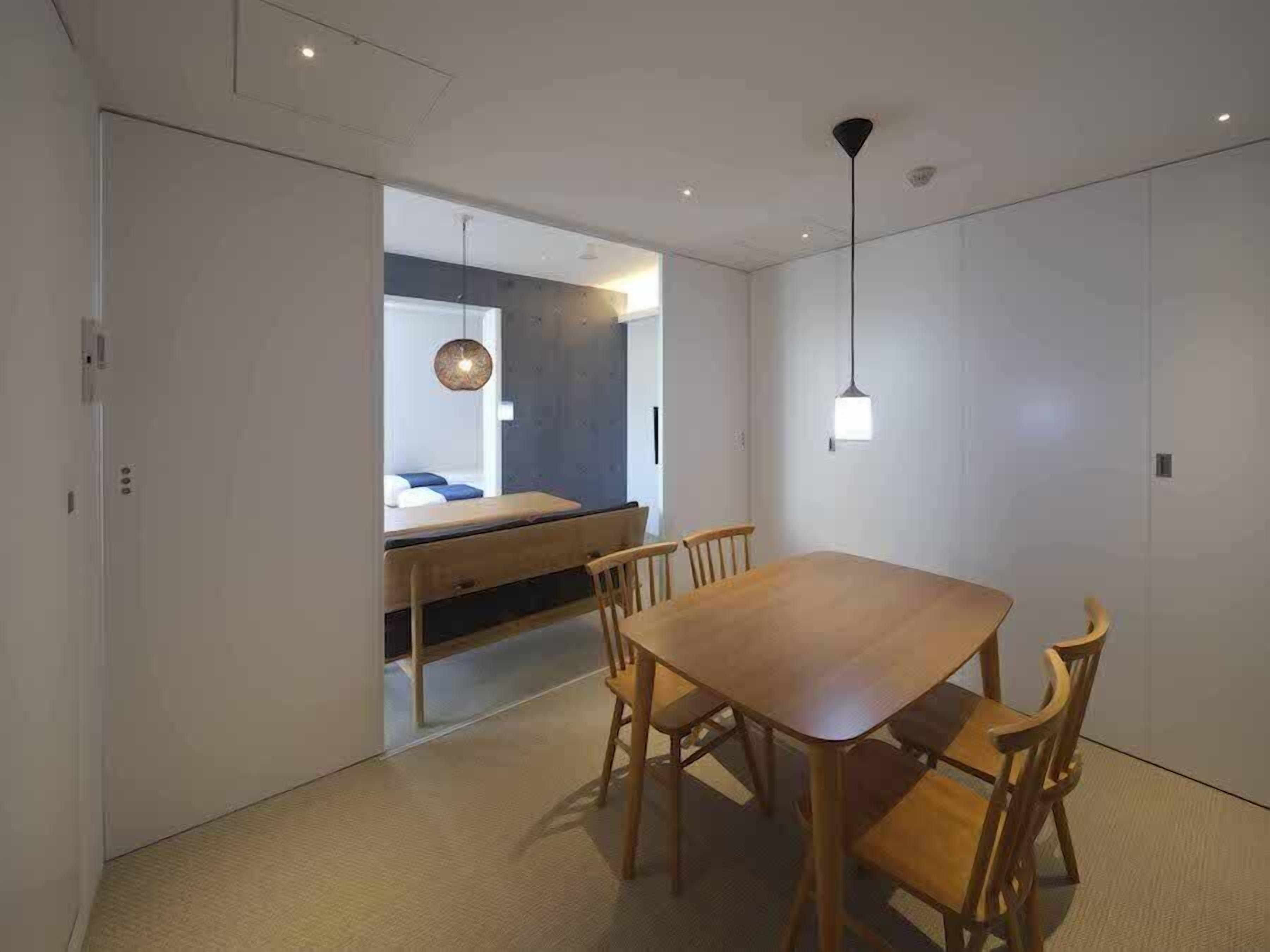 Un'area pranzo moderna con tavolo e sedie in legno, una lampada a sospensione e vista su una camera da letto con un letto e un lampadario.