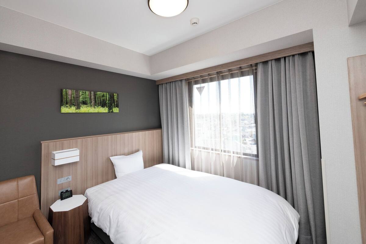 Ein Hotelzimmer mit Bett, Nachttisch, Stuhl, einem Bild und einem Fenster mit Vorhängen.