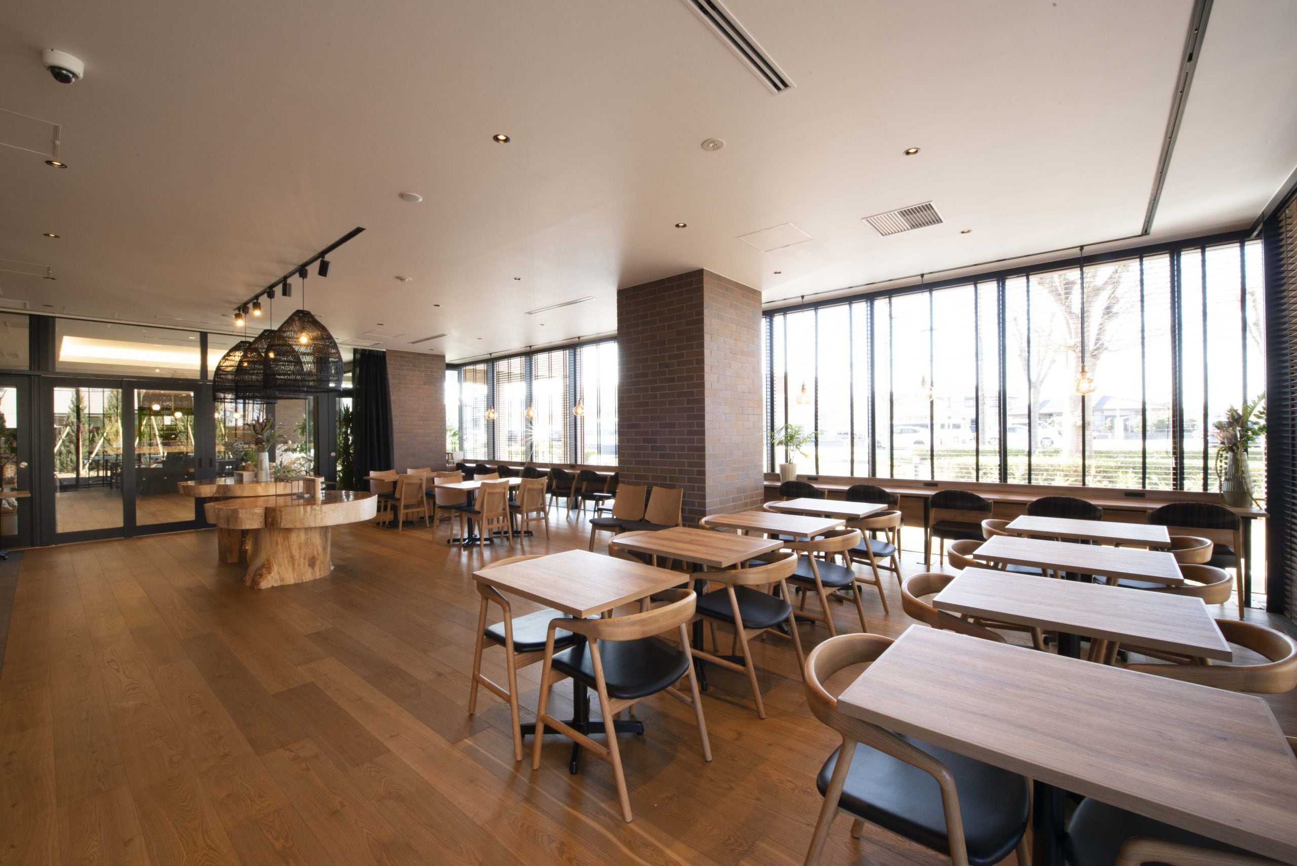 A spacious indoor area with multiple wooden tables and chairs, large windows providing natural light, and a brick column.