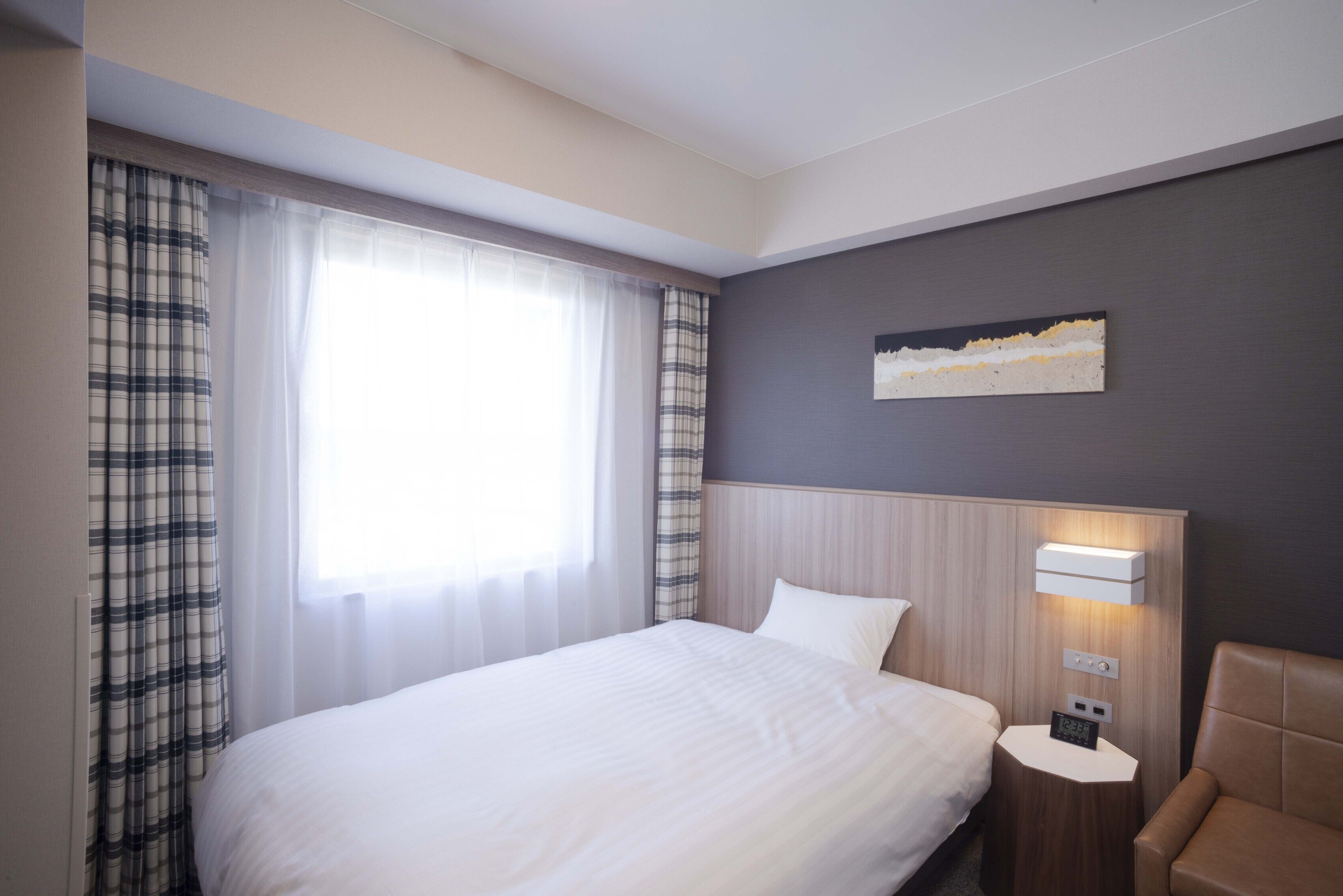 A hotel room with a bed, a nightstand, a lamp, a chair, and a painting on the wall.