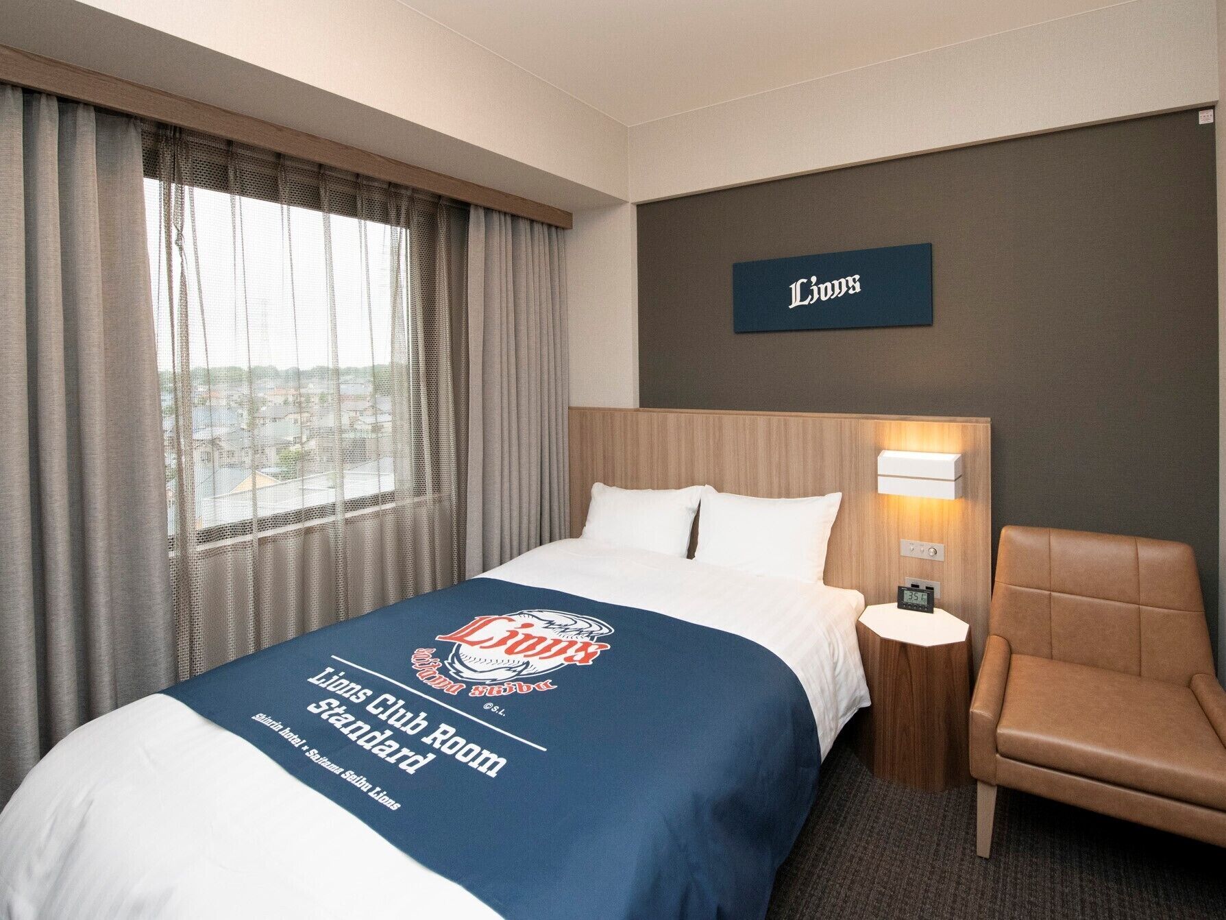 Habitación de hotel con cama, una silla, una mesita de noche, una lámpara y vista a la ciudad a través de la ventana.