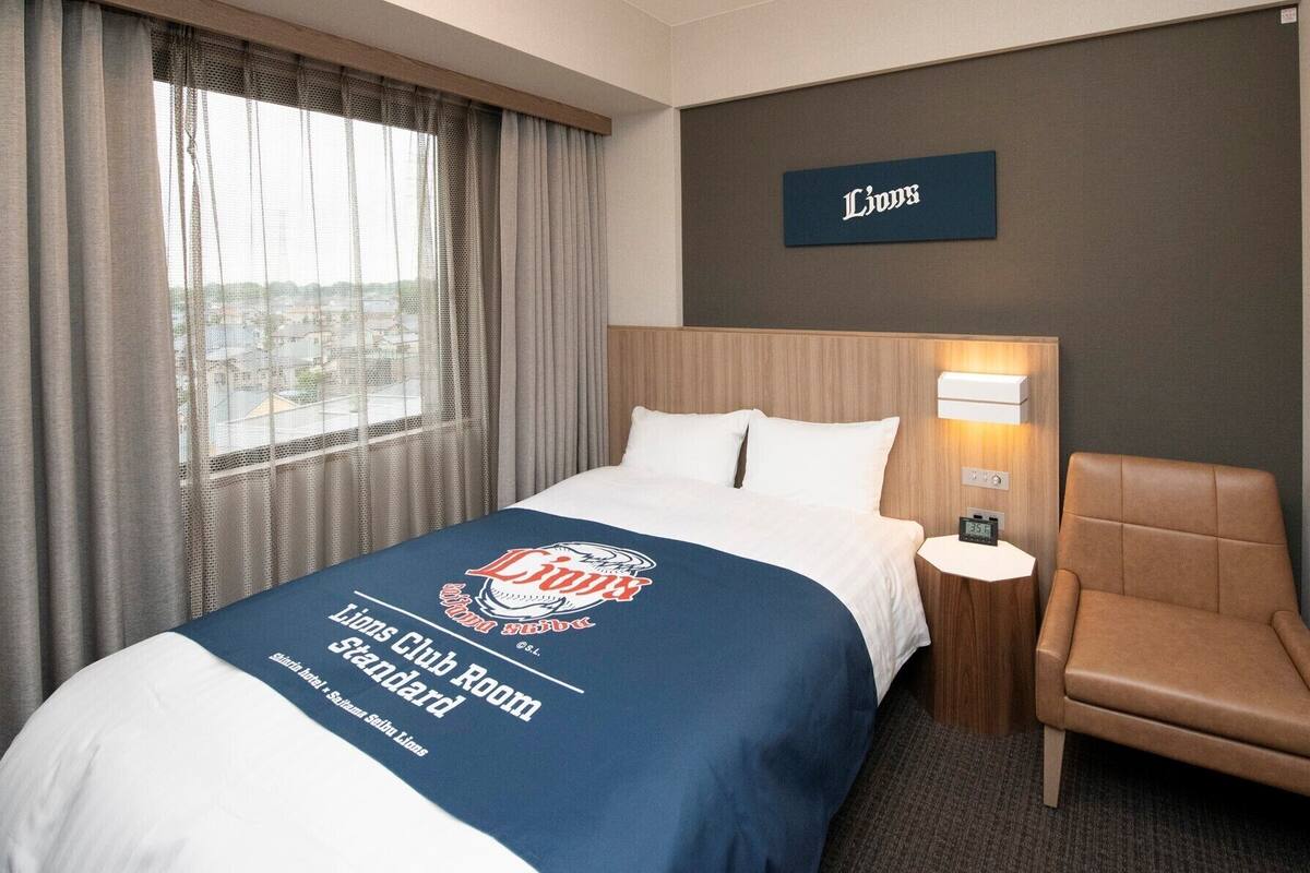 Ein Hotelzimmer mit Bett, Sessel, Nachttisch, Lampe und Blick auf die Stadt durch das Fenster.
