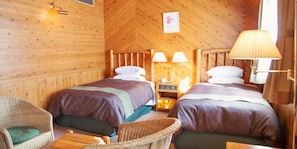 A wooden cabin room with two beds, a nightstand, a lamp, and wicker furniture. - Kibikougen Resort Hotel (Kibichuo)