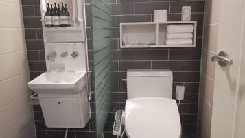 A bathroom with a white toilet, a sink, and a glass partition. There are bottles on the wall and a shelf with towels.