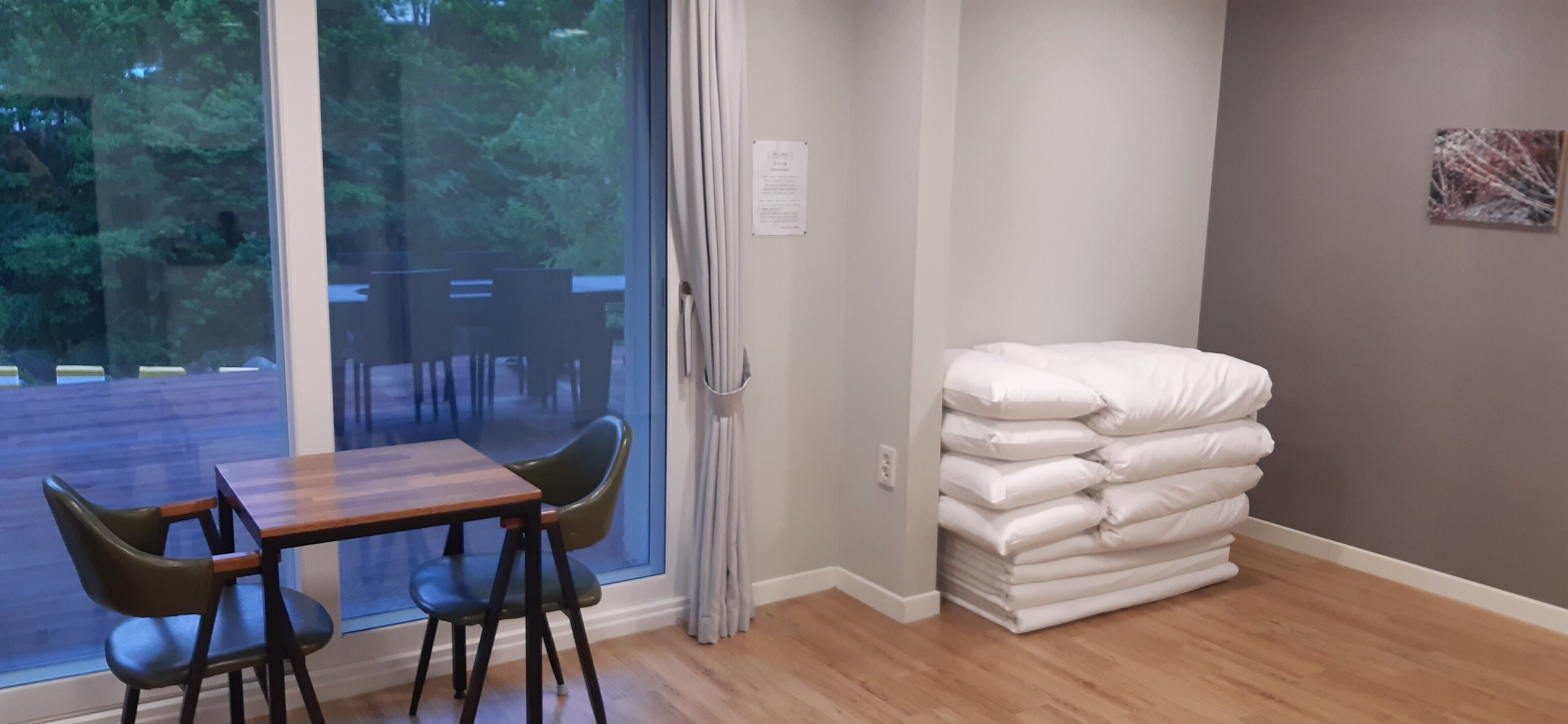 Una habitación con piso de madera, una mesa con dos sillas, un ventanal con cortinas y una pila de almohadas blancas apoyadas contra la pared.